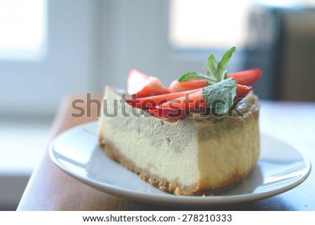 Slice of homemade cheesecake (with matcha green tea layer) decorated with fresh strawberries and mint on edge of kitchen table near window