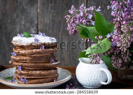 Stack of chocolate pancakes on wooden tray and lilac flowers, rustic wooden background