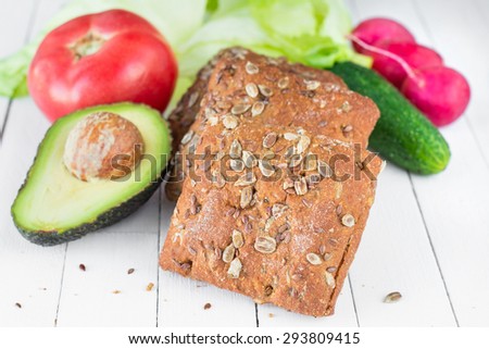 Fresh vegetables and whole wheat bread with seeds on white table, close up