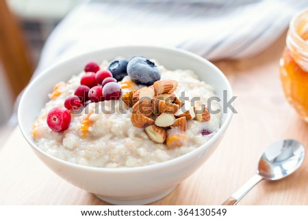 Oatmeal porridge with dried apricots, blueberries, cranberries and chopped almonds on bright wooden table. Jar of honey on background. Selective focus. Bright healthy breakfast image