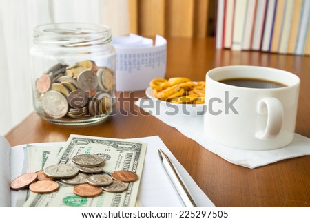 Many coins and banknotes place on notebook with a money saving jar, bills and coffee break on table, home finance concept