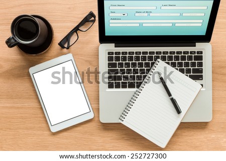 Open laptop shows blank information form and  digital tablet with isolate screen on wooden table. Blank diary on laptop, glasses and a cup of coffee on workspace. Top view image for mock up concept.