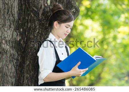 Asian student reading blue book, against green of summer park.
