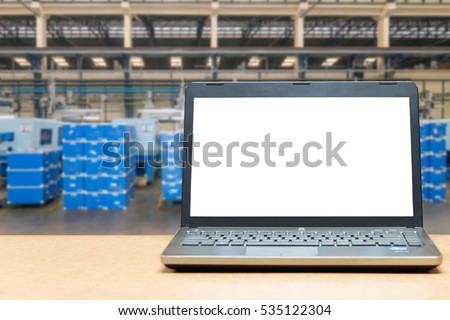 Laptop with blank screen on table with blur warehouse cargo in factory. Smart factory concept.