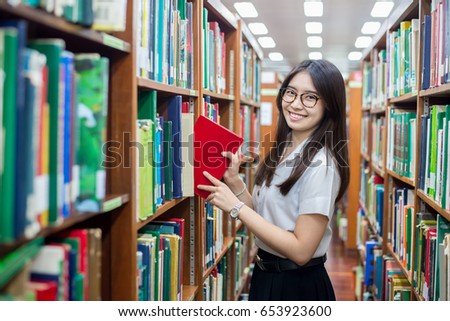 Cheerful female Asian student putting in order books returned after reading for literature lesson standing near bookshelves in modern interior library of university during break between lesson.