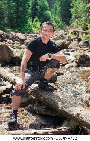 Man sitting on trunk on mountain trail. Mountain stream with stones and trees in background