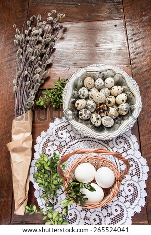 Easter composition of willow twigs with catkins, cress, quail eggs in metal dish and easter basket with eggs, set on old wooden table. Catkins are wrapped up in paper. Space for text at the top