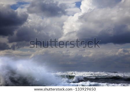 A giant wave raising above the sea\'s water surfaced, its surf scattered by the strong wind under the heavily overcast winter sky.