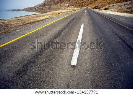 An empty road in the desert, shot with a very wide angle. The Sodom mountains are seen in the horizon, and the Dead sea is just to the left of the frame.