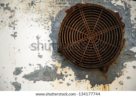 A vent in a metal wall, both in very bad shape with paint peeling off the wall and heavy corrosion on the vent cover.