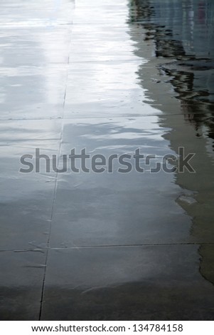 High angle vertical view of a wet concrete floor reflecting the changing luminosity of the inclement gray sky and a building, on a rainy winter day.