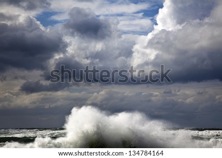 A giant wave raising above the sea\'s water surfaced, its surf scattered by the strong wind under the heavily overcast winter sky.
