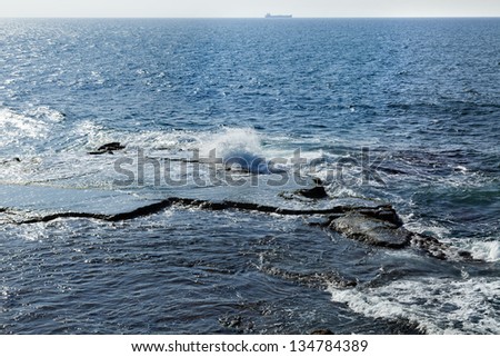 Waves crashing against a large flat rock in the Mediterranean sea. A large freighter breaking the horizon.