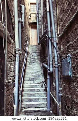 Narrow alley with pipes covered walls ends in a flight of stairs leading to an anonymous door.