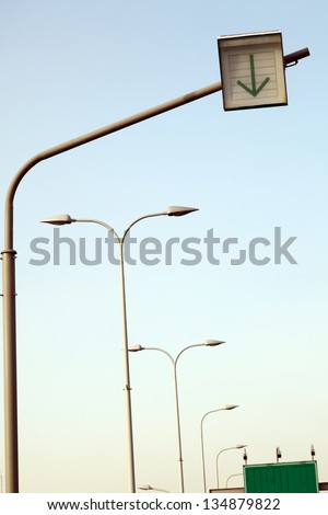 An overhead road sign followed by a row of street lights, with another green road sign in the back.