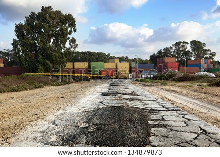 A badly patched broken asphalt road leads to a storage yard of cargo containers used for freight.