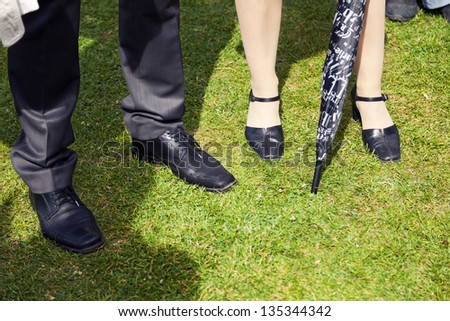 The legs of a businessman and a business woman with an umbrella, standing on green lawn outdoors in a sunny afternoon.