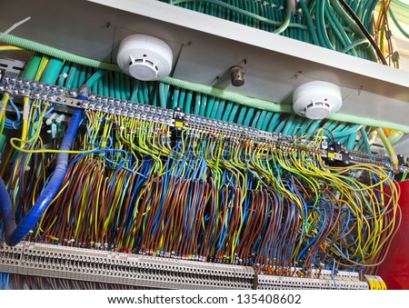 A large number of electric wires in various colors, distributing the electricity flow to the various locations in a large establishment. Above - two smoke detectors and a fire sprinkler.