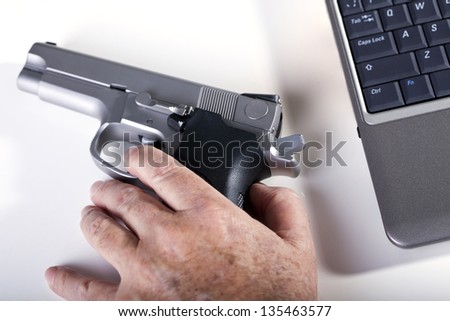 The left hand of a mature adult man resting on a 9mm handgun, and a laptop computer next to it.