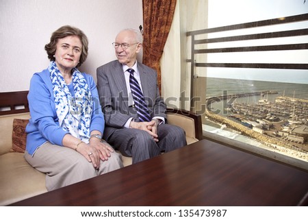 A high society senior couple (his 80's, her late 60's) sitting on a sofa. He's smiling and looking at her, and she's looking straight to the camera, with a slight smile on her face.
