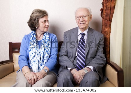 A high society senior couple (his 80's, her late 60's) sitting on a sofa. She's looking away to the right side of the frame, and he's looking straight to the camera, with a slight smile on his face.