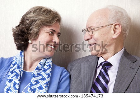 A high society senior couple (he's in his 80's, she's in her late 60's) sitting on a sofa looking at each other with very much love and joy.