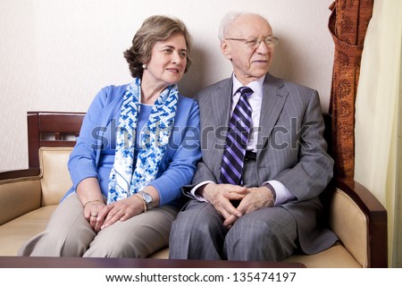 A high society senior couple (he's in his 80's, she's in her late 60's) sitting on a sofa looking away to the right side of the frame with very much love and joy.