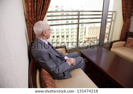 An elderly (in his 80\'s) business man wearing suit and tie sitting in a hotel\'s business lounge, looking through the window at the city view.