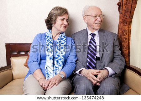 A high society senior couple (he\'s in his 80\'s, she\'s in her late 60\'s) sitting on a sofa looking away to the right side of the frame with very much love and joy.