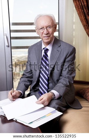 An elderly (80's) business man sitting in a hotel's business lounge, looking at camera with a slight smile on his face, in the middle of going over some papers after having coffee and cake.