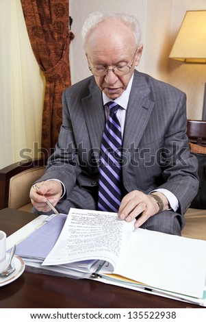 An elderly (in his 80's) business man wearing suit and tie sitting in a hotel's business lounge, going over some papers after having coffee and cake.