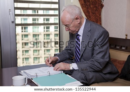 An elderly (in his 80\'s) business man wearing suit and tie sitting in a hotel\'s business lounge, going over some papers and writing notes after having coffee.