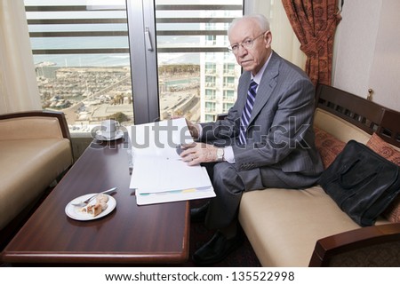 An elderly (in his 80\'s) business man wearing suit and tie sitting in a hotel\'s business lounge, looking at camera, as if disturbed in the middle of going over some papers after having coffee.