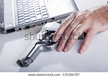 The left hand of a mature adult man resting on a 9mm handgun, and a defocused laptop computer in the background. Backlit.
