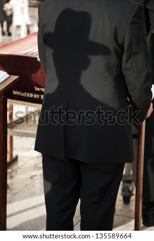 A shadow of an orthodox Jewish man casted on the back side of the suit of the orthodox Jewish man standing in front of him. Shot in the Wailing Wall in the old city of Jerusalem, Israel.