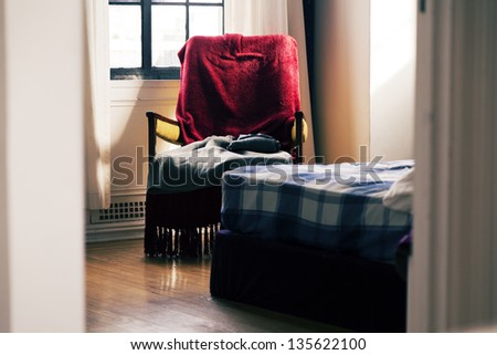A zoomed view at an armchair in a bedroom, partially hidden by the bed peeking in. Shot through the door of the room, creating a voyeuristic feeling.