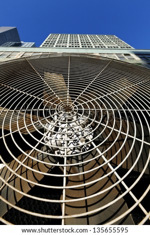 Low & wide angle close-up of an HVAC air-conditioner outdoor unit\'s ventilator, with a midtown NYC skyscraper rising above it on the background of clear blue sky