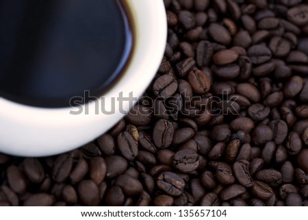 Part of a cup filled with black coffee on background of coffee beans. Shot from high angle, focus on the beans in the background.