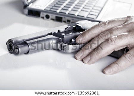 The left hand of a mature adult man resting on a 9mm handgun, and a laptop computer in the background. Back lit.
