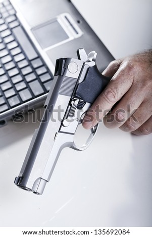 The left hand of a mature adult man holding a 9mm handgun, and a laptop computer in the background. Back lit. Shallow depth of field.