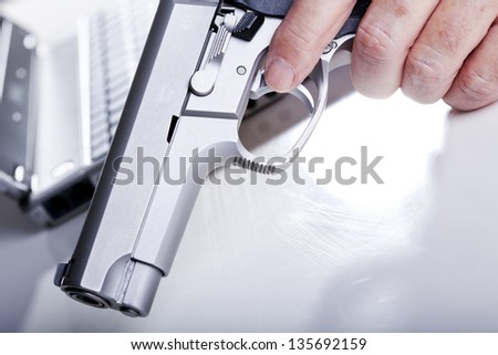 Extreme close up of a left hand of a mature adult man holding a 9mm handgun with his finger on the trigger, and a laptop computer in the background. Back lit. Shallow depth of field.