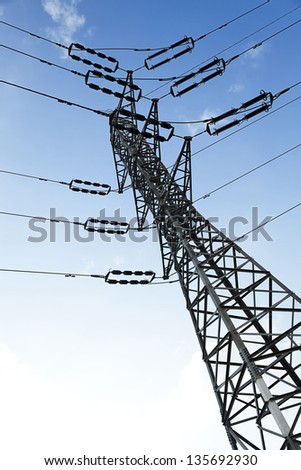 Low angle tilted shot of a high-voltage electricity pylon and power lines, on the background of the blue & white cloudy sky.
