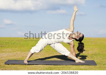 An adult woman standing on a black yoga mattress in the Utthita Trikonasana (aka Extended Triangle) pose, on a green lawn with cloudy blue sky in the background.
