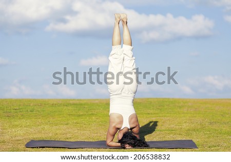 An adult woman standing on her head on a black yoga mattress in the Salamba Sirsasana (aka Supported Headstand) pose, on a green lawn with cloudy blue sky in the background.