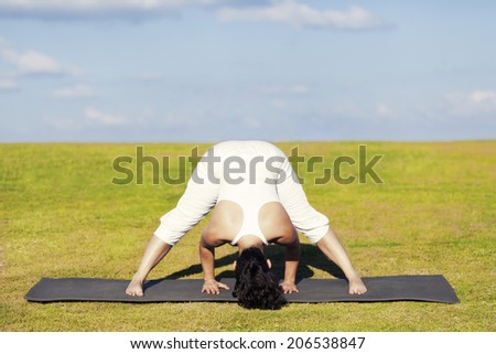An adult woman standing on a black yoga mattress in the Prasarita Padottanasana (aka Wide-Legged Standing Forward Bend) pose, on a green lawn with cloudy blue sky in the background.
