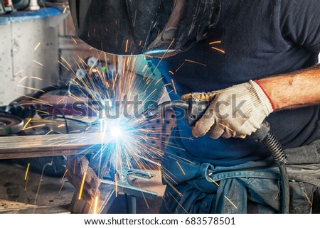 A man welder in a black T-shirt, construction gloves and a welding mask hard work and  welds with a welding machine metal in workshop, close up