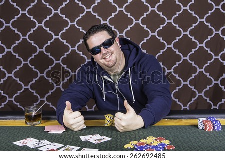A man sitting at a poker table wearing a hoodie gambling playing cards against a brown background