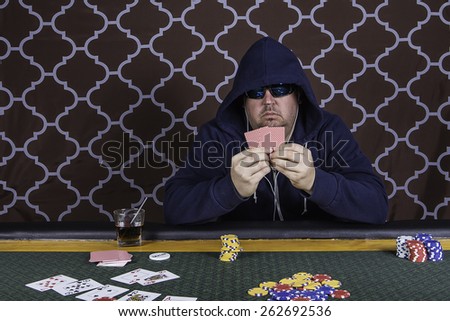 A man sitting at a poker table wearing a hoodie gambling playing cards against a brown background