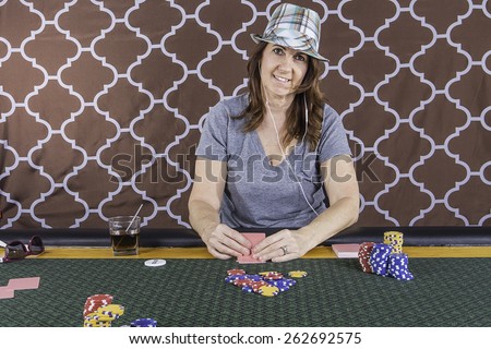 A woman sitting at a poker table wearing a hat playing cards with a brown background