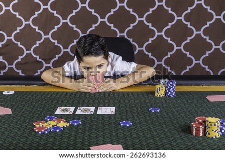 A young boy sitting at a poker table gambling playing cards with a brown background